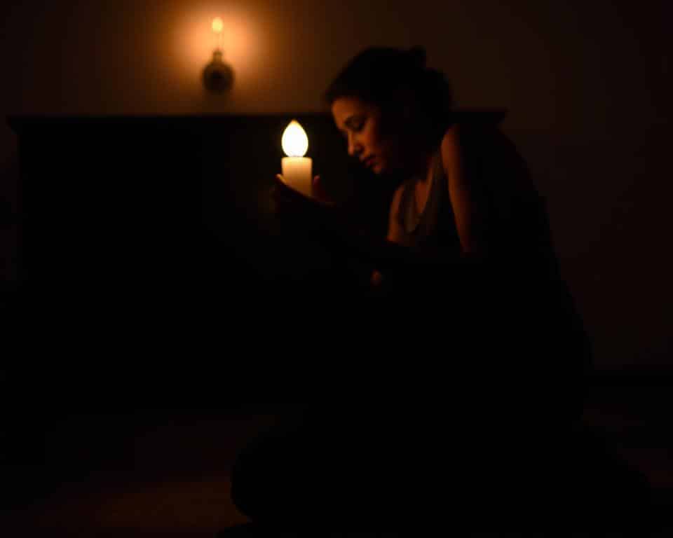 A person kneeling in a dimly lit room, their hands clasped in prayer, face illuminated by the soft glow of a single candle. The background is blurred, creating a sense of solitude and focus. The camera captures the scene from a low angle, emphasizing the reverence and intimacy of the moment. The lighting is warm and inviting, casting gentle shadows that add depth and texture to the image. The atmosphere is one of tranquility and devotion, conveying the sense of a personal, spiritual connection.